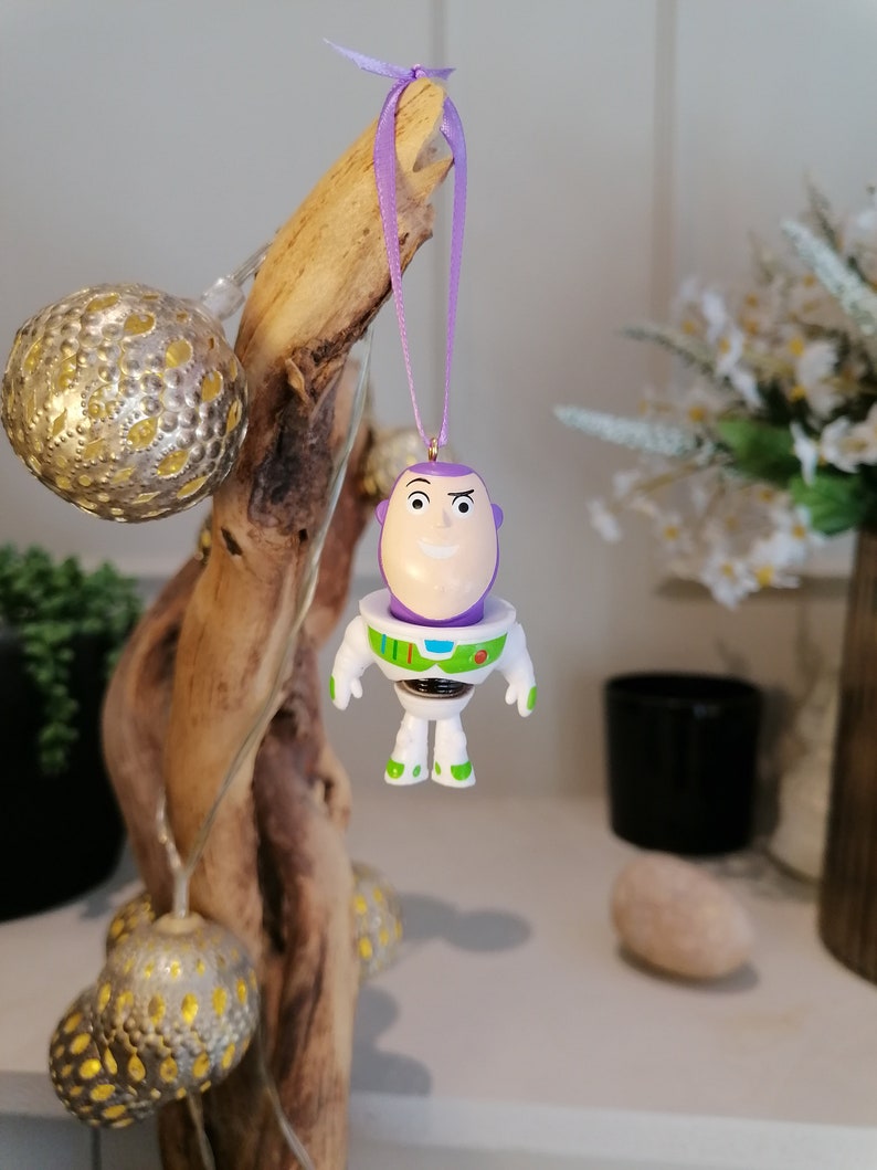 Puede incluir: Adorno navide&ntilde;o de Buzz Lightyear morado y blanco con una cinta morada. El adorno est&aacute; colgado de una rama.