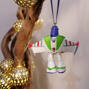 Pu&ograve; includere: Un ornamento natalizio Buzz Lightyear bianco, verde e blu con accenti rossi. L'ornamento &egrave; appeso a un nastro blu su un ramo con luci.