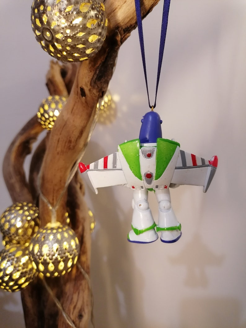 Pu&ograve; includere: Un ornamento natalizio Buzz Lightyear bianco, verde e blu con accenti rossi. L'ornamento &egrave; appeso a un nastro blu su un ramo con luci.