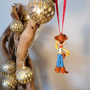 Pu&ograve; includere: Un ornamento di plastica rosso e bianco di Woody di Toy Story. Woody indossa il suo cappello da cowboy e il gilet caratteristici. Sta tenendo il suo lazo nella mano destra. L'ornamento &egrave; appeso a un nastro rosso.