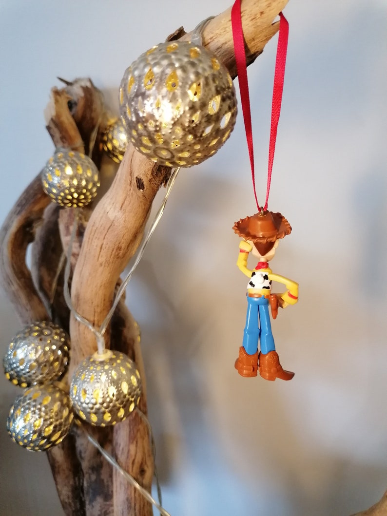Pu&ograve; includere: Un ornamento di plastica rosso e bianco di Woody di Toy Story. Woody indossa il suo cappello da cowboy e il gilet caratteristici. Sta tenendo il suo lazo nella mano destra. L'ornamento &egrave; appeso a un nastro rosso.