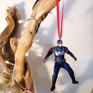 May include: A blue and red superhero action figure ornament hanging from a branch with white lights. The figure is holding a red, white, and blue shield.