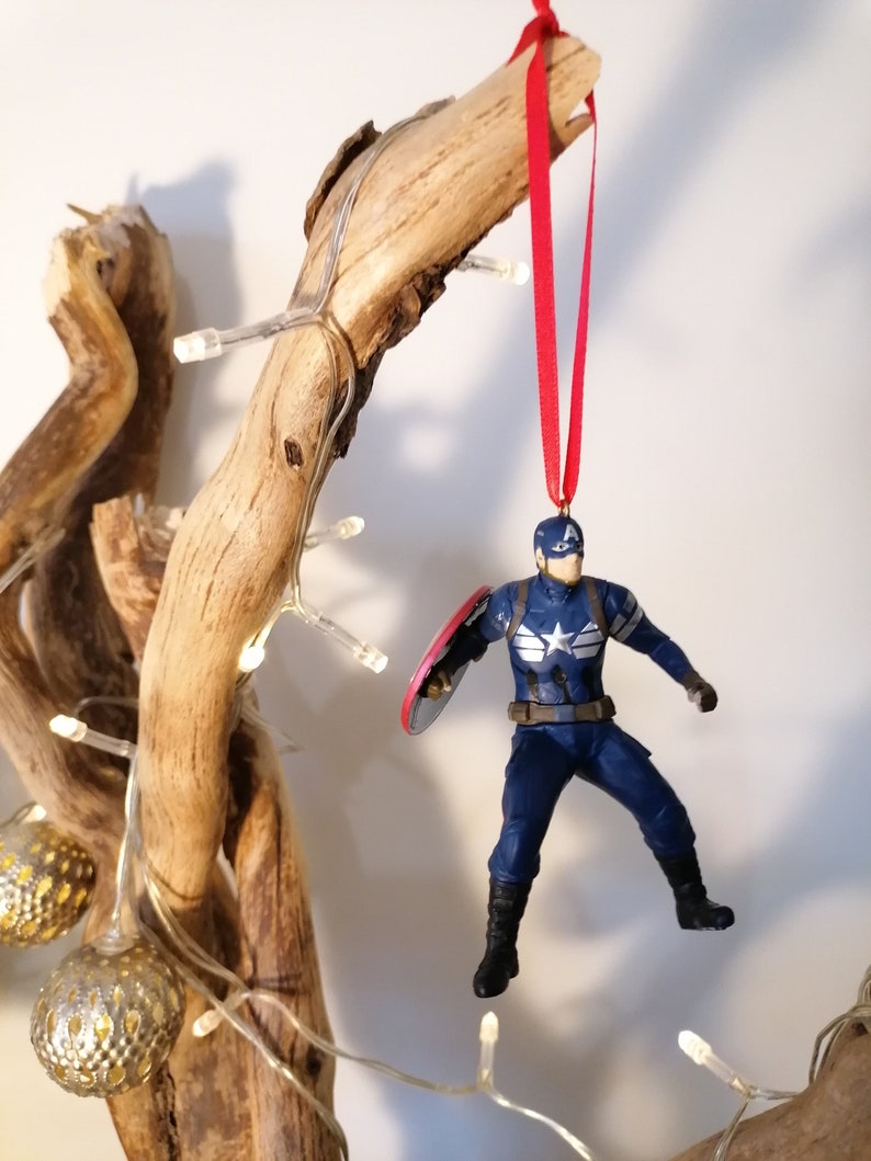 May include: A blue and red superhero action figure ornament hanging from a branch with white lights. The figure is holding a red, white, and blue shield.