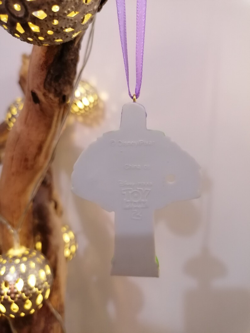 Puede incluir: Adorno de cer&aacute;mica blanca con forma de Buzz Lightyear de la franquicia Toy Story. El adorno tiene una cinta morada. En la parte posterior del adorno se encuentra el logotipo de Disney Pixar y las palabras "Toy Story" y "25th Anniversary".