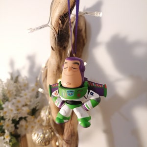K&ouml;nnte beinhalten: Ein lila und gr&uuml;ner Weihnachtsbaumschmuck in Form von Buzz Lightyear aus der Toy Story Filmreihe. Der Schmuck hat einen lila Helm, einen gr&uuml;nen K&ouml;rper und wei&szlig;e Arme und Beine. Der Schmuck h&auml;ngt an einem lila Band.