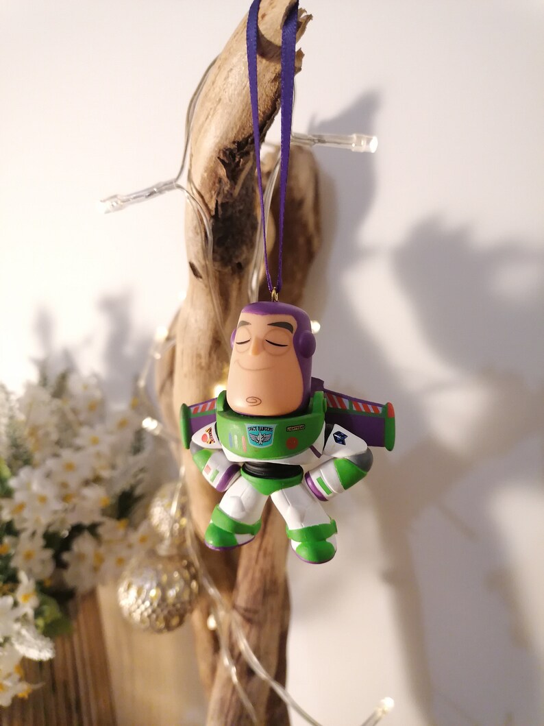 K&ouml;nnte beinhalten: Ein lila und gr&uuml;ner Weihnachtsbaumschmuck in Form von Buzz Lightyear aus der Toy Story Filmreihe. Der Schmuck hat einen lila Helm, einen gr&uuml;nen K&ouml;rper und wei&szlig;e Arme und Beine. Der Schmuck h&auml;ngt an einem lila Band.