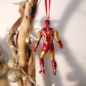 May include: Red and gold Iron Man Christmas ornament hanging from a branch. The ornament is a detailed figurine of Iron Man in his red and gold suit.