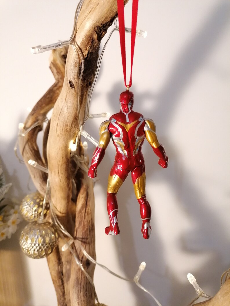 May include: Red and gold Iron Man Christmas ornament hanging from a branch. The ornament is a detailed figurine of Iron Man in his red and gold suit.