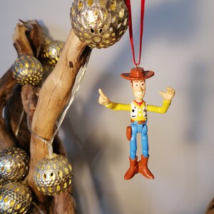 May include: A plastic toy figurine of Woody from Toy Story hanging from a red ribbon. Woody is wearing his signature cowboy hat, vest, and blue jeans. He is holding his right arm up in the air, as if he is waving. The figurine is hanging from a branch with silver, decorative, round ornaments.