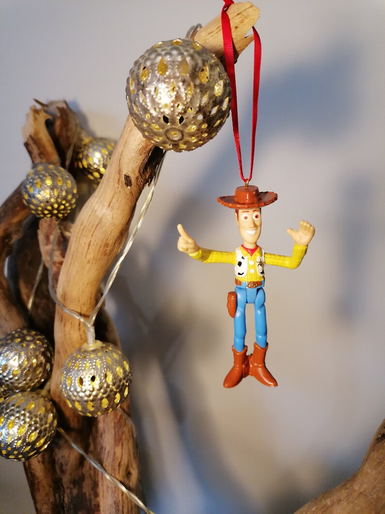 May include: A plastic toy figurine of Woody from Toy Story hanging from a red ribbon. Woody is wearing his signature cowboy hat, vest, and blue jeans. He is holding his right arm up in the air, as if he is waving. The figurine is hanging from a branch with silver, decorative, round ornaments.
