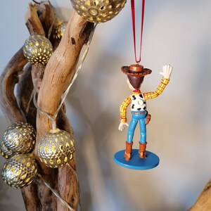 May include: A red string hangs from a wooden branch with a silver ornament. A plastic toy figurine of Woody from Toy Story is hanging from the string. Woody is wearing his signature cowboy hat, vest, and jeans. He is waving with his right hand. The figurine is on a blue base.