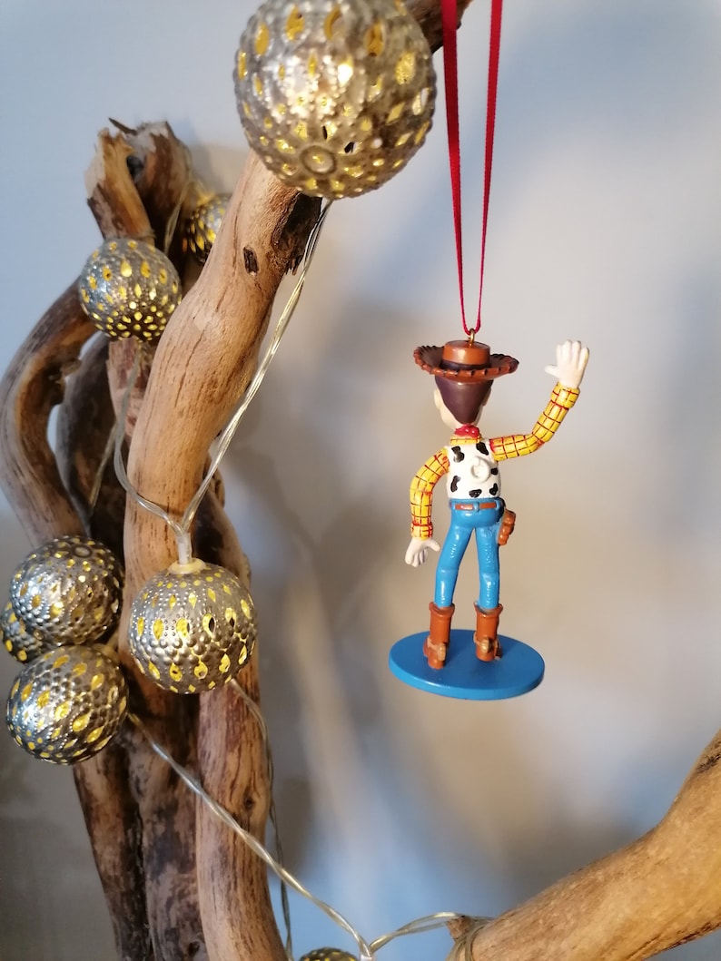 May include: A red string hangs from a wooden branch with a silver ornament. A plastic toy figurine of Woody from Toy Story is hanging from the string. Woody is wearing his signature cowboy hat, vest, and jeans. He is waving with his right hand. The figurine is on a blue base.