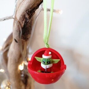 Pu&ograve; includere: Un ornamento natalizio rosso con un personaggio verde e marrone che indossa un cappello da Babbo Natale. L'ornamento &egrave; appeso a un nastro verde.