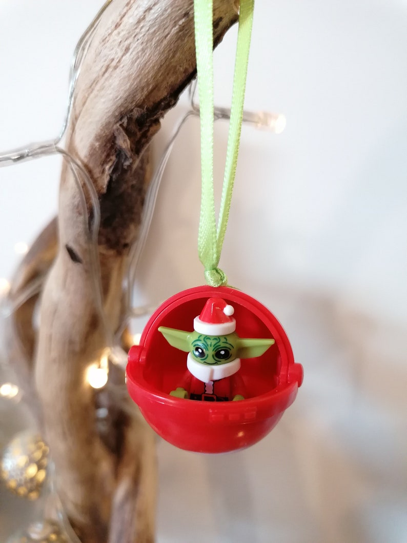 Pu&ograve; includere: Un ornamento natalizio rosso con un personaggio verde e marrone che indossa un cappello da Babbo Natale. L'ornamento &egrave; appeso a un nastro verde.