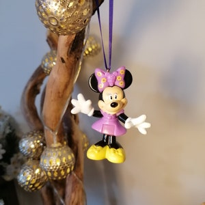 May include: Purple Minnie Mouse Christmas ornament with a yellow bow and black ears. The ornament is hanging from a purple ribbon on a wooden branch.