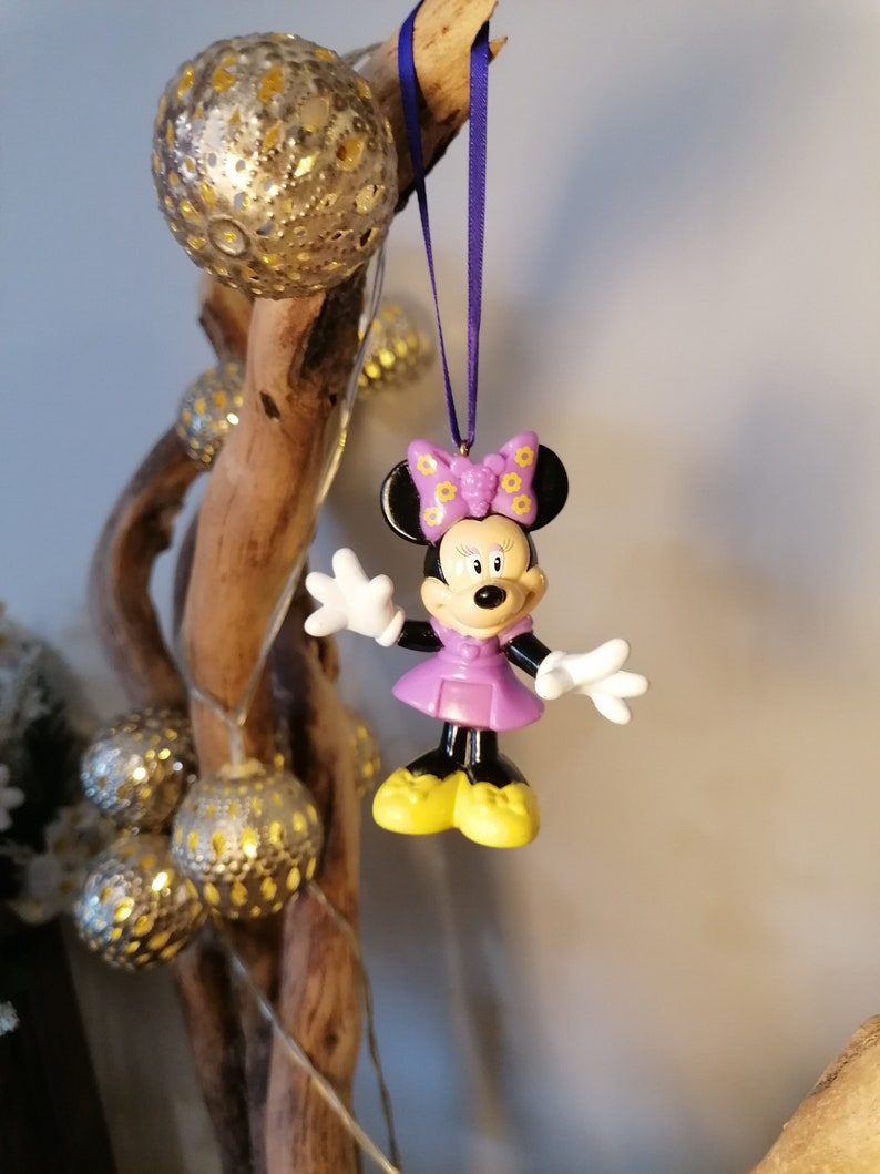 May include: Purple Minnie Mouse Christmas ornament with a yellow bow and black ears. The ornament is hanging from a purple ribbon on a wooden branch.