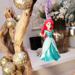 May include: A plastic figurine of Ariel, the Little Mermaid, wearing a teal gown and a crown. She has long red hair and is hanging from a purple ribbon.