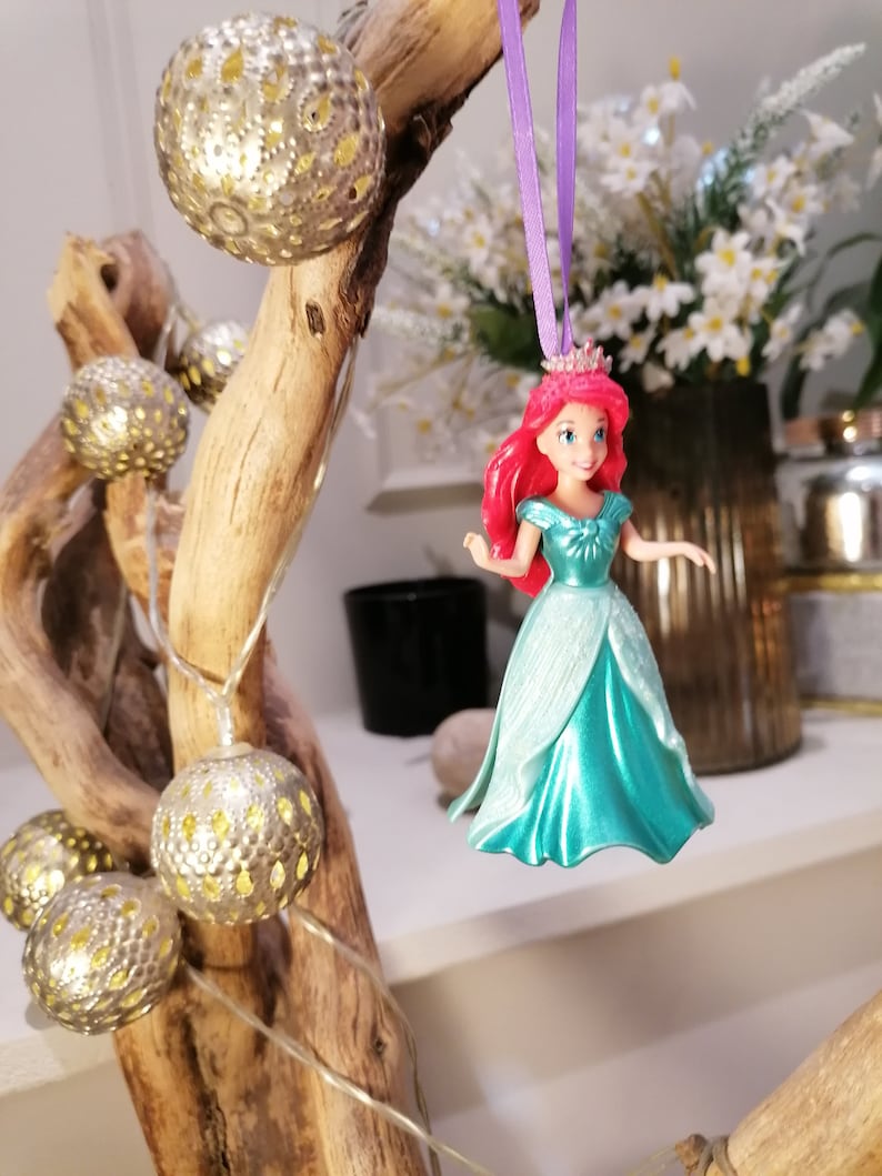 May include: A plastic figurine of Ariel, the Little Mermaid, wearing a teal gown and a crown. She has long red hair and is hanging from a purple ribbon.