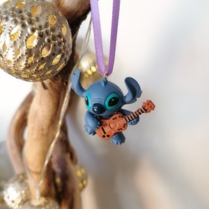 Op de afbeelding: Een blauwe Stitch-figuur ornament dat een bruine ukelele bespeelt, hangend aan een paars lint.
