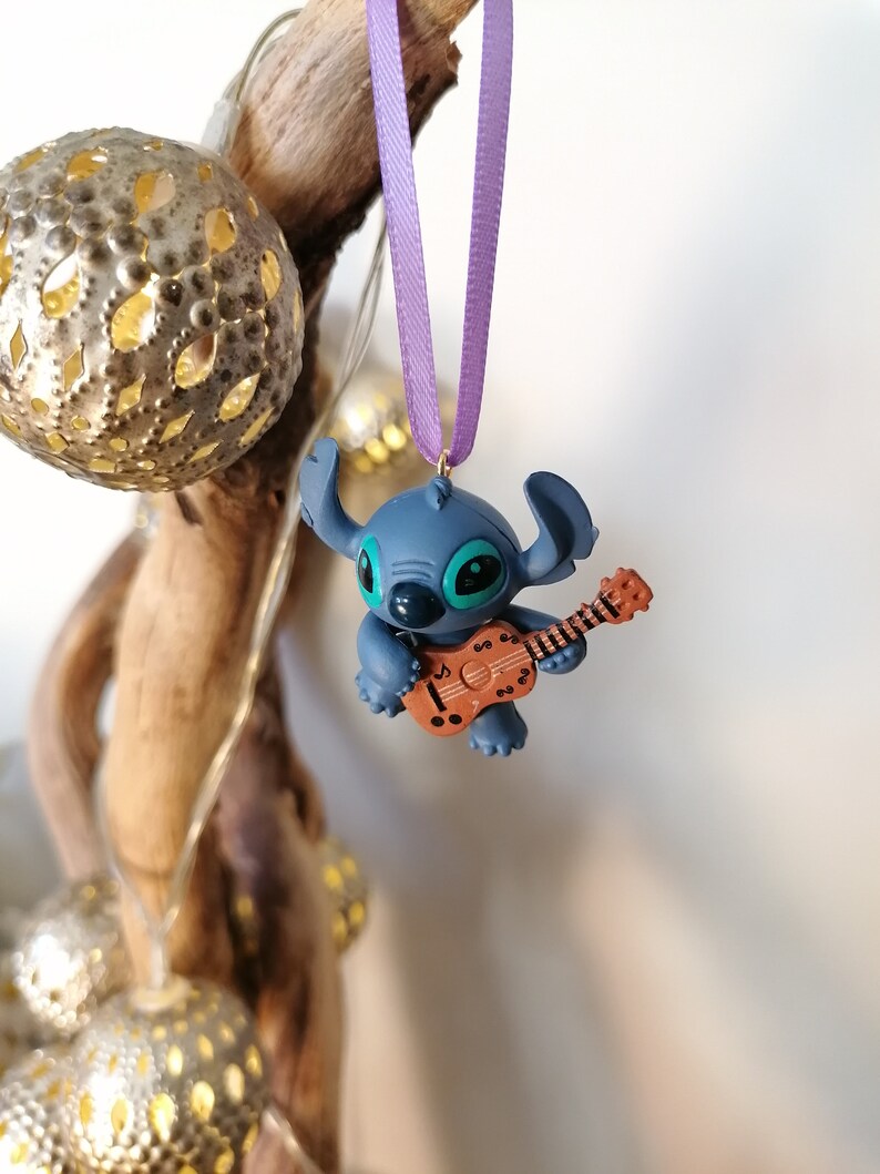 Op de afbeelding: Een blauwe Stitch-figuur ornament dat een bruine ukelele bespeelt, hangend aan een paars lint.