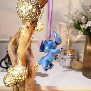 Pode incluir: Um ornamento azul do Stitch pendurado em uma fita roxa. O ornamento &eacute; feito de pl&aacute;stico e tem um acabamento brilhante. O ornamento est&aacute; pendurado em um galho com luzes de corda.