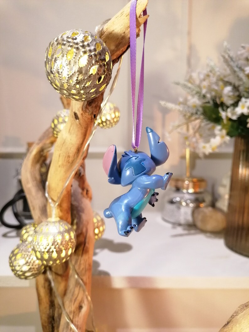Pode incluir: Um ornamento azul do Stitch pendurado em uma fita roxa. O ornamento &eacute; feito de pl&aacute;stico e tem um acabamento brilhante. O ornamento est&aacute; pendurado em um galho com luzes de corda.
