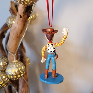 May include: A red and white checkered shirt with a brown cowboy hat and blue jeans. The figure is holding a gun and has a brown belt. The figure is a Christmas ornament.