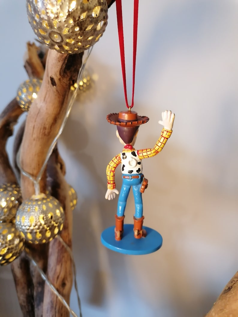 May include: A red and white checkered shirt with a brown cowboy hat and blue jeans. The figure is holding a gun and has a brown belt. The figure is a Christmas ornament.
