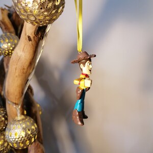 May include: A Woody action figure ornament, featuring a brown cowboy hat, yellow shirt, blue pants, and brown boots. The ornament is suspended by a yellow ribbon. Decorative silver globe lights and a wooden branch are in the background.