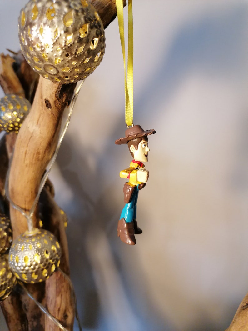 May include: A Woody action figure ornament, featuring a brown cowboy hat, yellow shirt, blue pants, and brown boots. The ornament is suspended by a yellow ribbon. Decorative silver globe lights and a wooden branch are in the background.