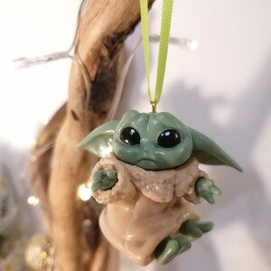 May include: A green and brown Christmas ornament featuring a character from the Star Wars franchise, The Mandalorian. The ornament is hanging from a green ribbon.