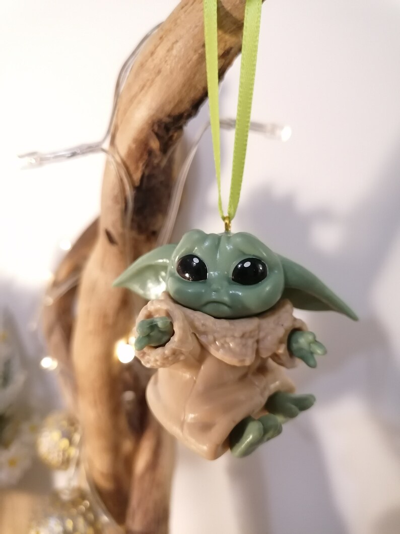 May include: A green and brown Christmas ornament featuring a character from the Star Wars franchise, The Mandalorian. The ornament is hanging from a green ribbon.
