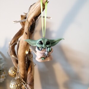 May include: Green and pink Christmas ornament of a character from the Star Wars franchise, The Mandalorian. The ornament is hanging from a branch with a green ribbon.