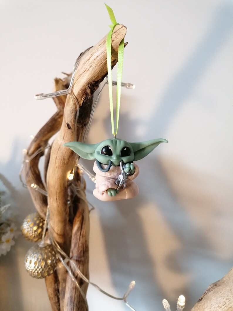 May include: Green and pink Christmas ornament of a character from the Star Wars franchise, The Mandalorian. The ornament is hanging from a branch with a green ribbon.