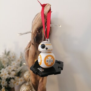 May include: A white and orange Star Wars BB-8 character ornament with a black base. The ornament is hanging from a red ribbon on a piece of driftwood.
