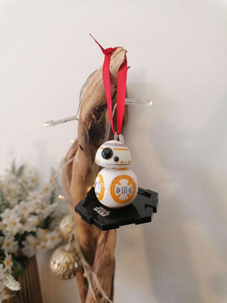 May include: A white and orange Star Wars BB-8 character ornament with a black base. The ornament is hanging from a red ribbon on a piece of driftwood.