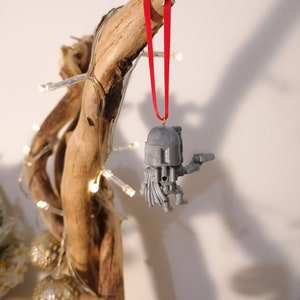 May include: A grey plastic Star Wars bauble depicting a character wearing a helmet and holding a blaster. The bauble is hanging from a red ribbon on a branch.