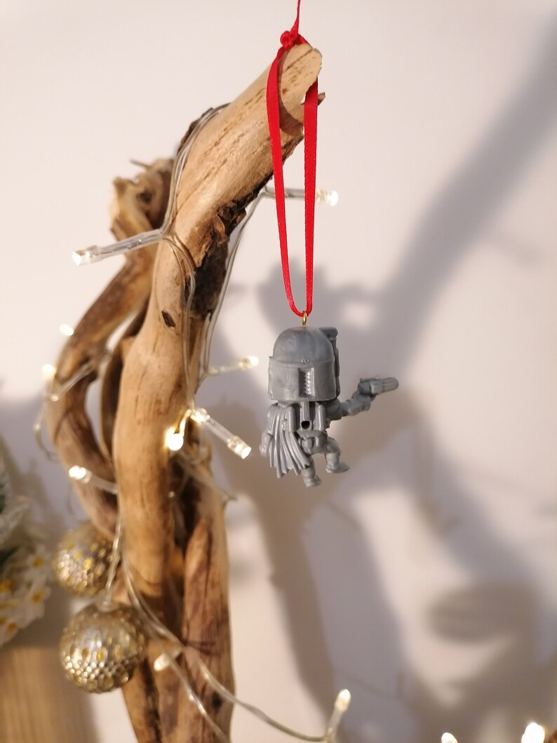 May include: A grey plastic Star Wars bauble depicting a character wearing a helmet and holding a blaster. The bauble is hanging from a red ribbon on a branch.