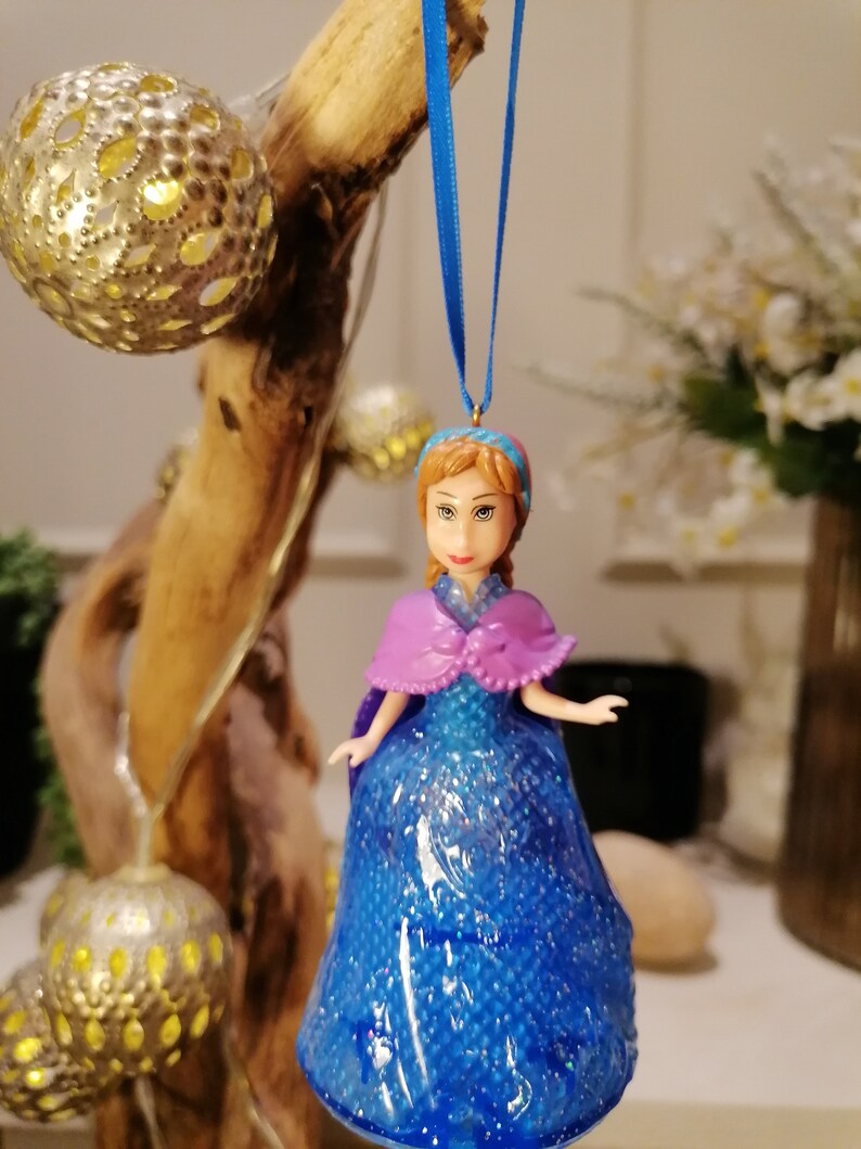 Puede incluir: Un adorno de vestido azul y morado con purpurina que presenta una mu&ntilde;eca con cabello casta&ntilde;o y un sombrero azul y blanco. El adorno est&aacute; colgado de una cinta azul.