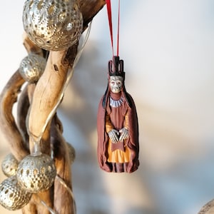May include: A brown and gold Christmas ornament depicting a figure wearing a crown and robe. The ornament is hanging from a red ribbon.