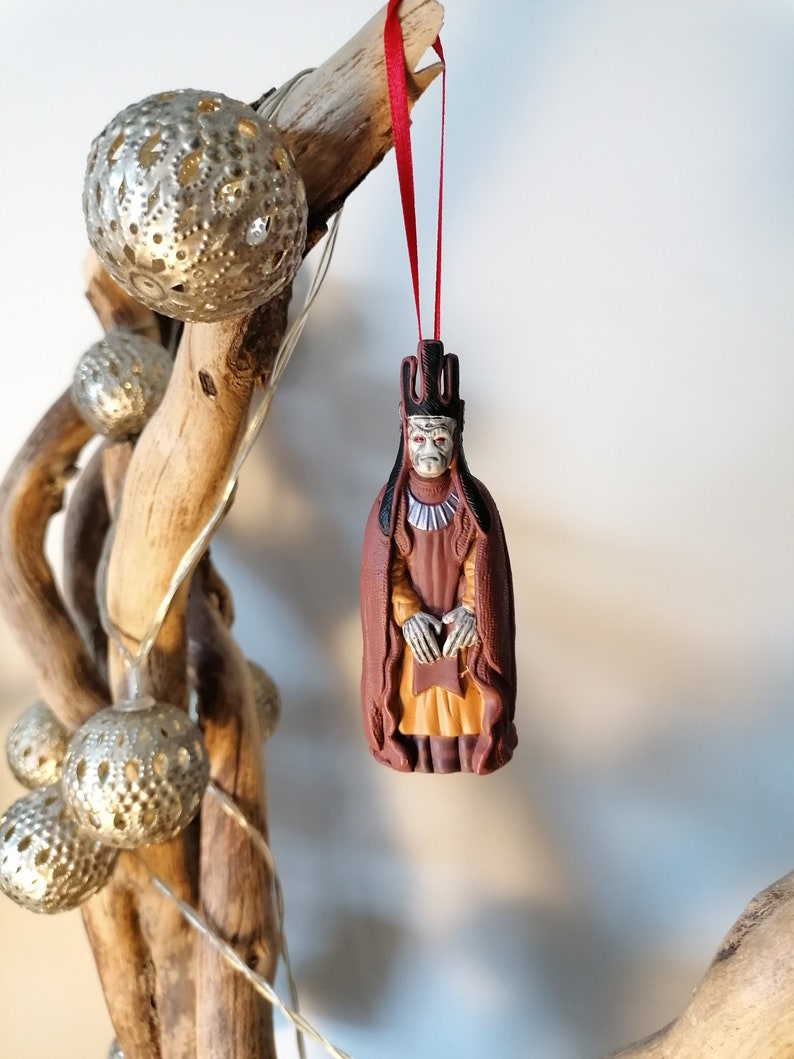 May include: A brown and gold Christmas ornament depicting a figure wearing a crown and robe. The ornament is hanging from a red ribbon.