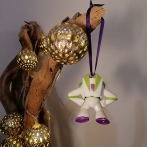 May include: A white and green plastic toy astronaut ornament with purple accents hanging from a purple ribbon on a branch with string lights. The lights are in gold and white spheres with a cut-out design.