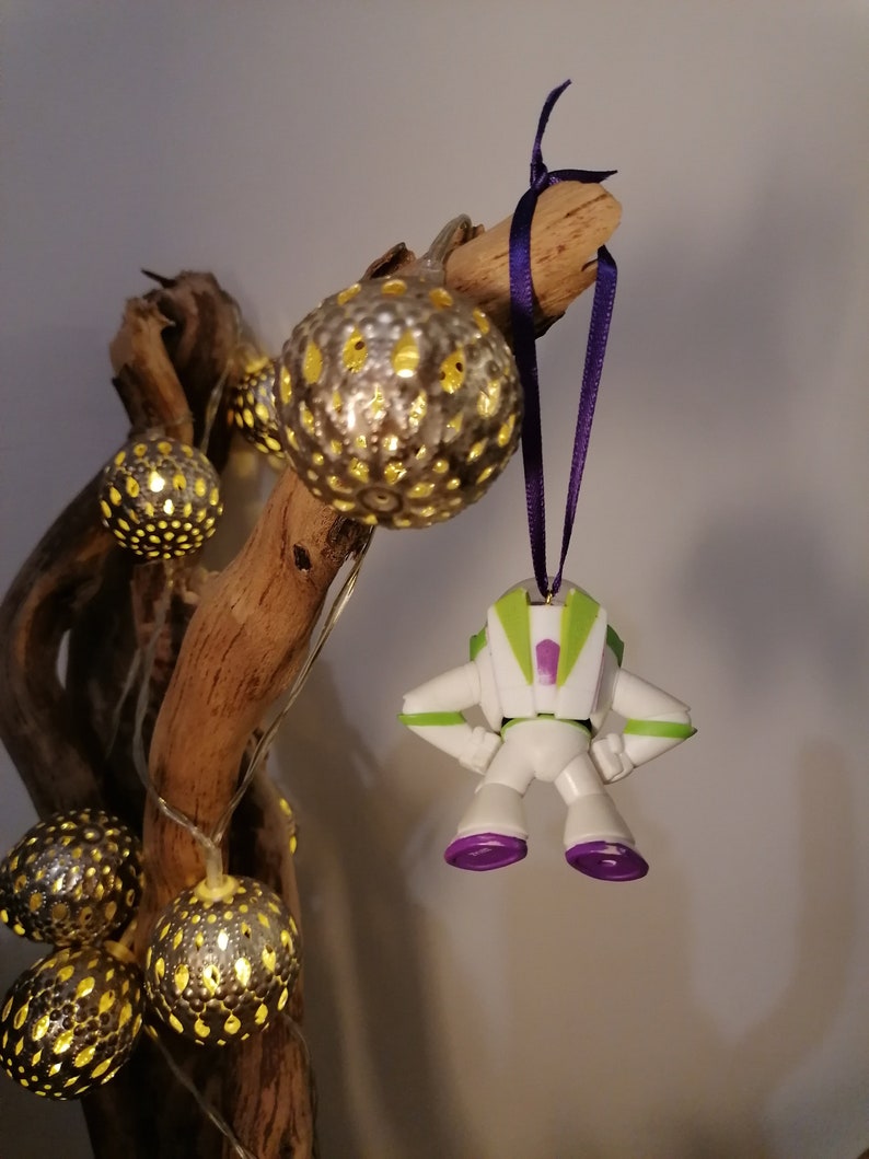 May include: A white and green plastic toy astronaut ornament with purple accents hanging from a purple ribbon on a branch with string lights. The lights are in gold and white spheres with a cut-out design.