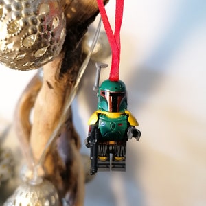 May include: A green and yellow Lego Star Wars Boba Fett ornament hanging from a red ribbon. The ornament is a miniature version of the character Boba Fett from the Star Wars franchise.