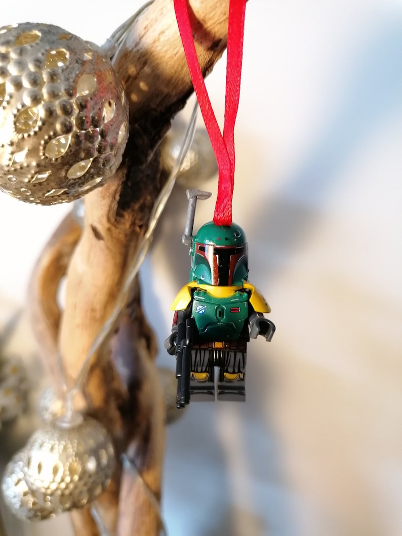 May include: A green and yellow Lego Star Wars Boba Fett ornament hanging from a red ribbon. The ornament is a miniature version of the character Boba Fett from the Star Wars franchise.