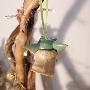 May include: A green and brown Yoda ornament hanging from a branch with a green ribbon. The ornament is a popular character from the Star Wars franchise.