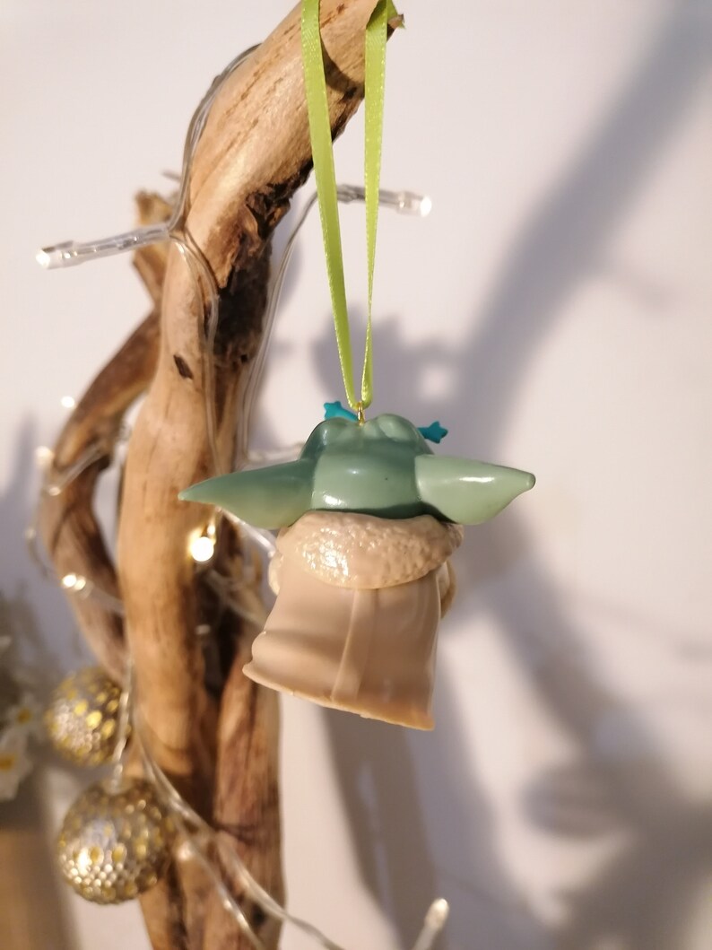 May include: A green and brown Yoda ornament hanging from a branch with a green ribbon. The ornament is a popular character from the Star Wars franchise.