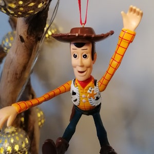 May include: A red-stringed Christmas ornament featuring a plastic figurine of Woody from Toy Story. Woody is wearing his signature brown cowboy hat, yellow shirt, and blue jeans. He is waving with his right arm and has a friendly expression on his face.