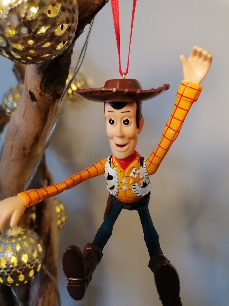 May include: A red-stringed Christmas ornament featuring a plastic figurine of Woody from Toy Story. Woody is wearing his signature brown cowboy hat, yellow shirt, and blue jeans. He is waving with his right arm and has a friendly expression on his face.