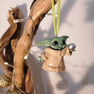 May include: A green and brown Christmas ornament shaped like a character from the Star Wars franchise. The ornament is holding a silver ball in its hand.